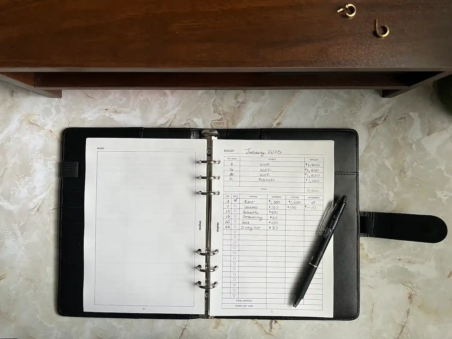 An open A5 planner sits on a desk displaying a filled-in budgeting sheet with income and expenses alongside a Pilot FriXion erasable clicker pen.