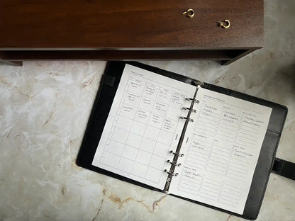 An open planner with a meal planner and grocery list page visible, placed on a marble-textured surface. Categories include meal alternatives and pantry essentials.