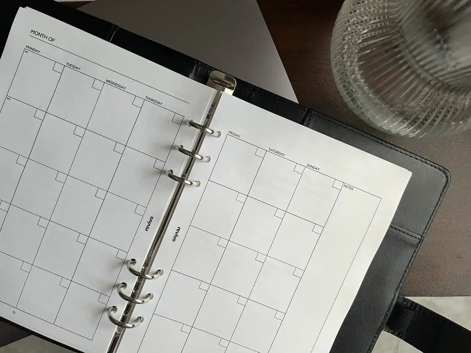 A monthly planner layout with a clean grid calendar design, displayed in a sleek black leather binder alongside a textured glass vase, perfect for organizing schedules and appointments.